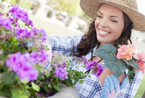 A woman in a gardening hat and blue gloves is tending to a variety of vibrant flowering plants in a garden. The garden features a neatly maintained lawn area with lush green grass, bordered by flower beds containing purple and pink blooms. In the background, there are mature trees and a paved pathway, suggesting a well-designed landscaped outdoor space typical of residential gardens in the Addiscombe area. The woman is smiling while carefully handling the flowers, indicating active gardening or planting. The natural daylight illuminates the scene, highlighting the vivid colours of the flowers and the healthy condition of the garden. This setting demonstrates detailed garden elements such as the soft grass surface, lush foliage, and neatly arranged flower beds, reflecting professional gardening and lawn care practices that Lawn Mowing Addiscombe offers in the local community.